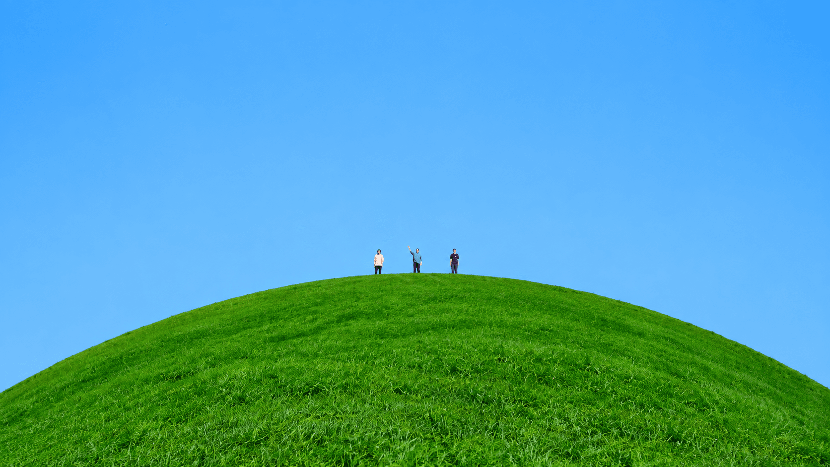 Three friends standing on top of a round green hill under a clear blue sky